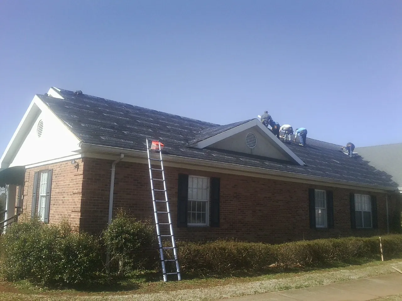 Crew tearing shingles off a brick ranch roof with a ladder set — Emergency Roof Repair work by our crew in Wind Ridge