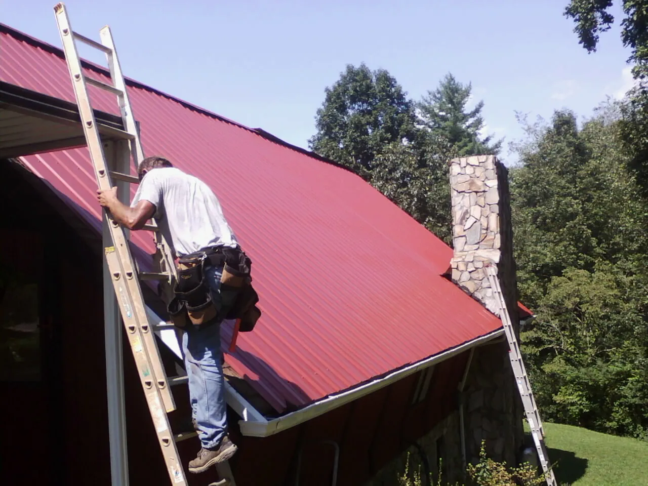 Roofer on a ladder beside a red metal roof and stone chimney — Soffit & Fascia Repair by Roofing Brothers in Wind Ridge